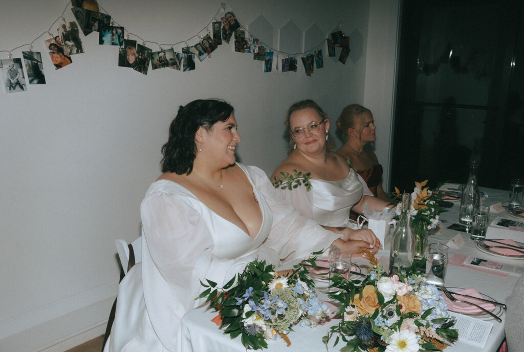 Two brides smiling as they are seated in Juliet Somerville for their restaurant wedding reception.