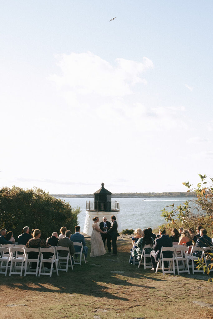 Wide shot of guests as they witness bride and groom getting married. Scenery includes the Castle Hill Lighthouse, lawn, and ocean. Photo by Rhode Tripper Photography