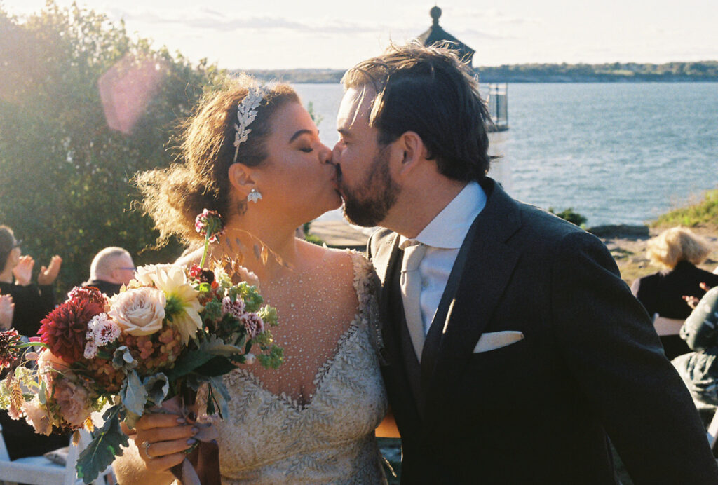 Bride and groom kiss as they come back down the aisle together. Sun flares in the left corner and the lighthouse peeks from behind the groom. Photo by Rhode Tripper Photography