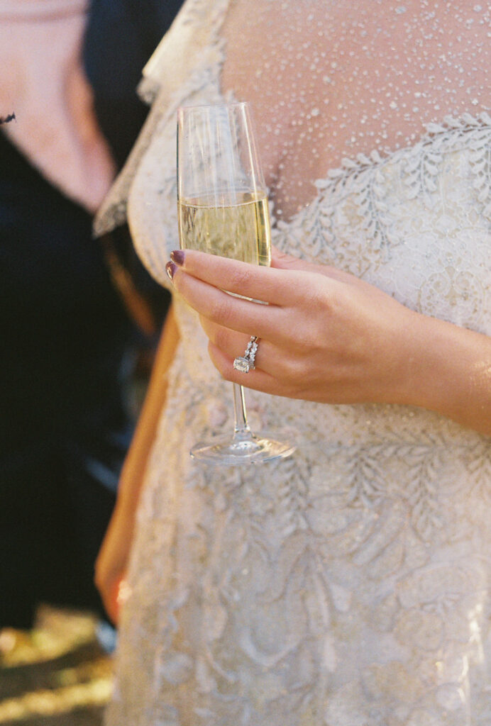 Close up of the bride's ring finger with her new wedding band and engagement ring showcased. Bride is holding a glass of champagne and wearing a lace wedding dress.