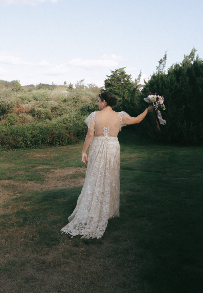 Back view of the bride in her wedding dress holding her bouquet in the air. Surrounded by lush green grass and bushes at Castle Hill.