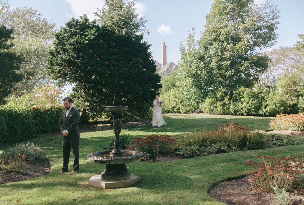 Groom waits with hands folded for his bride in the garden at Castle Hill Inn. Bride approaches with bouquet in hand in the background. Photo by Rhode Tripper Photography