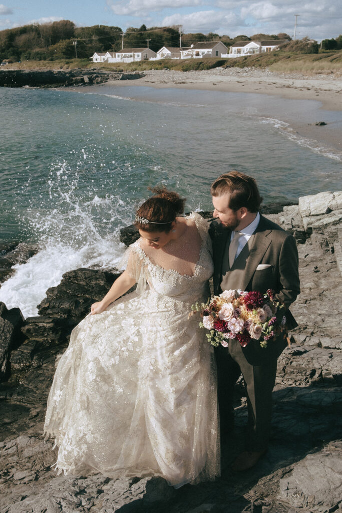 Bride and groom look towards the train of the bride's sparkly, lace wedding dress. Bride holds her train out atop the rocks as the waves crash beneath them. Castle Hill Inn beach cottages create the background. Photo by Rhode Tripper Photography