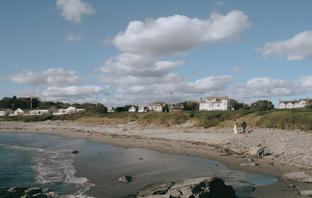 Wide shot of the beach at Castle Hill Inn as a couple walks along the shore in their wedding attire. Photo by Rhode Tripper Photography