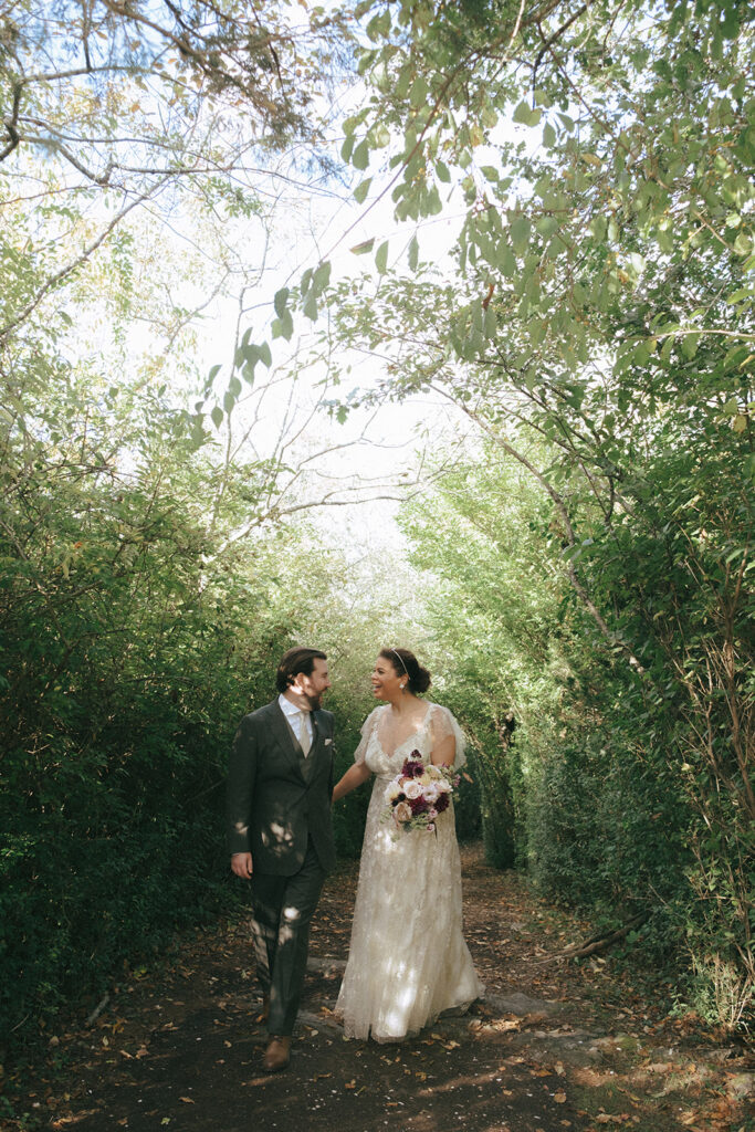 Green trees create a natural arch over the bride and groom as they walk and laugh together along a pathway at Castle Hill Inn. Photo by Rhode Tripper Photography