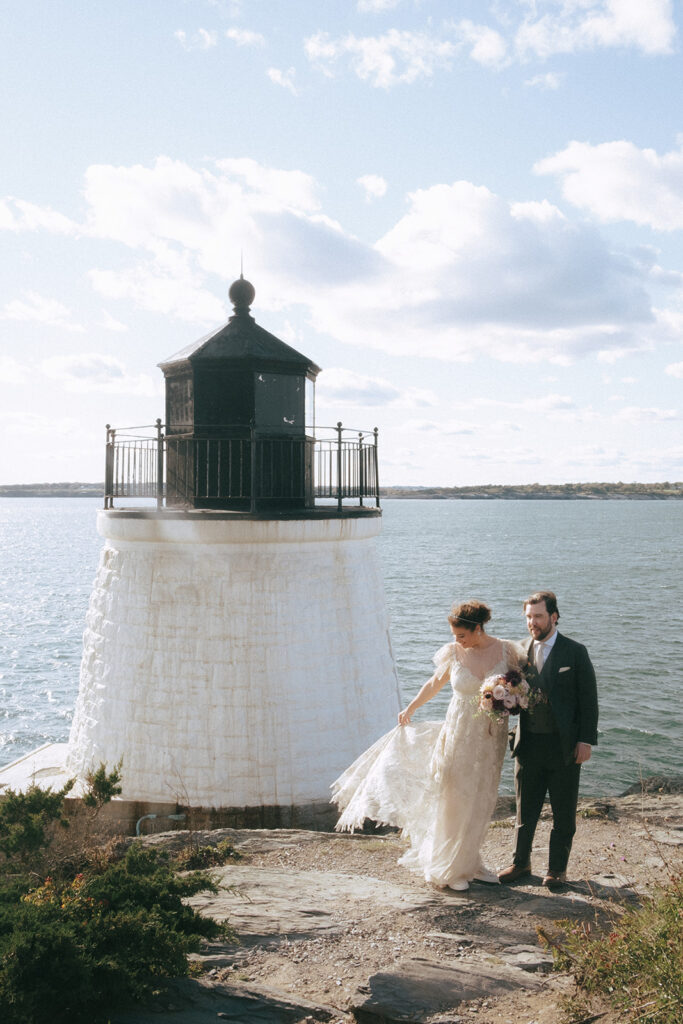 Bride throws lace wedding dress train into the wind while standing next to her groom. Scenery includes the cliffs and Castle Hill Lighthouse with Narragansett Bay in the background. Photo by Rhode Tripper Photography