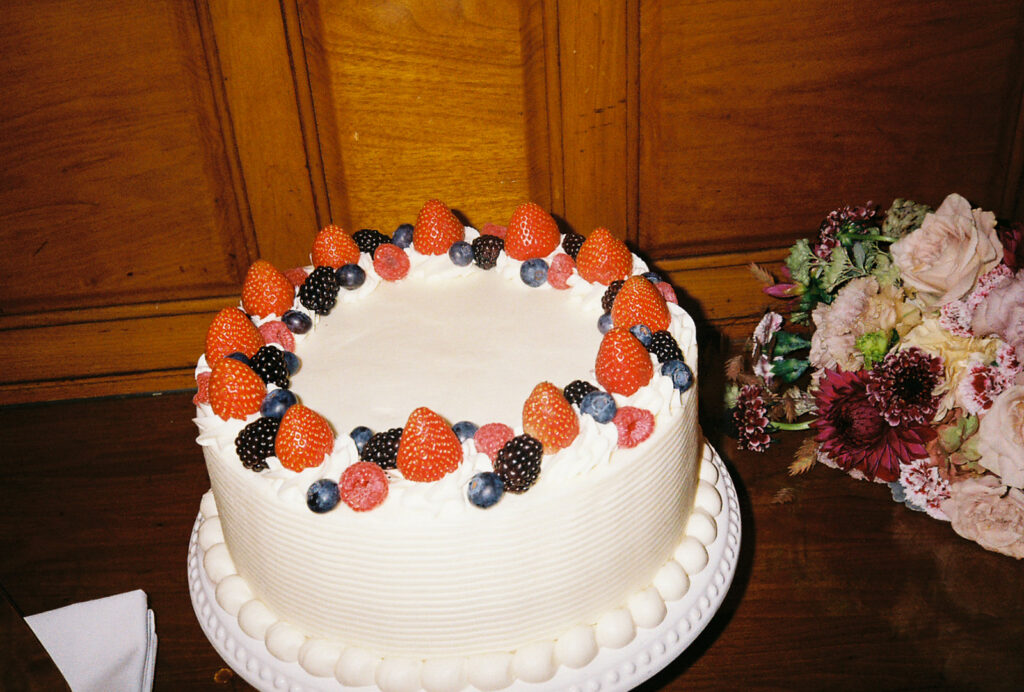 Film photo shows a white single tier wedding cake with berries atop it and the bride's bouquet in the background. Photo by Rhode Tripper Photography