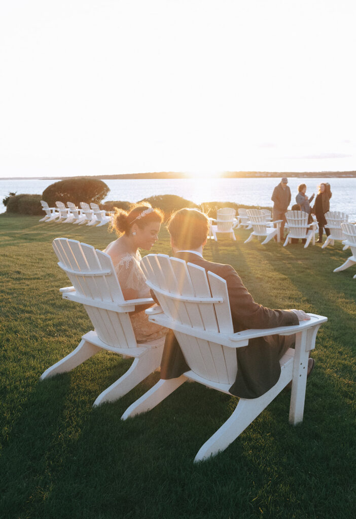 Couple wearing wedding attire looks out towards the sunset over Narragansett bay. Seated in Adirondack chairs on the Castle Hill Lawn with a sun flare in the background.