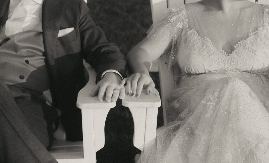 Close up in black and white of bride and groom's hands while seated in Adirondack chairs on the lawn of Castle Hill. Photo by Rhode Tripper Photography