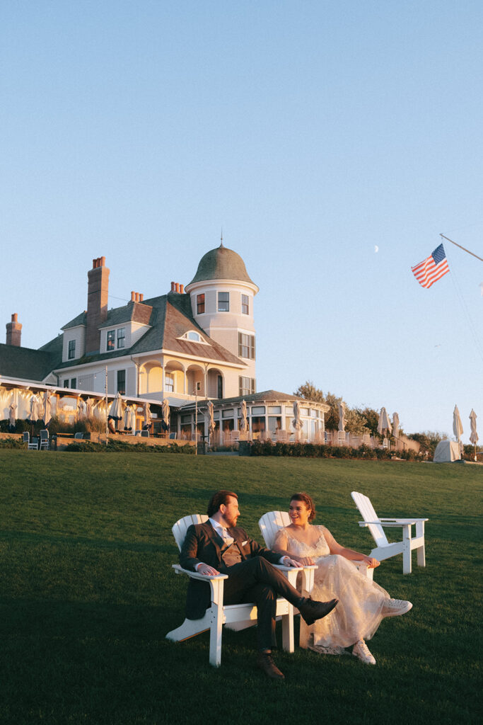 Bride and groom seated in white Adirondack chairs. In the background sits the historic mansion of Castle Hill Inn. Light is golden and pink showcasing the beautiful sunset hour. Photo by Rhode Tripper Photography