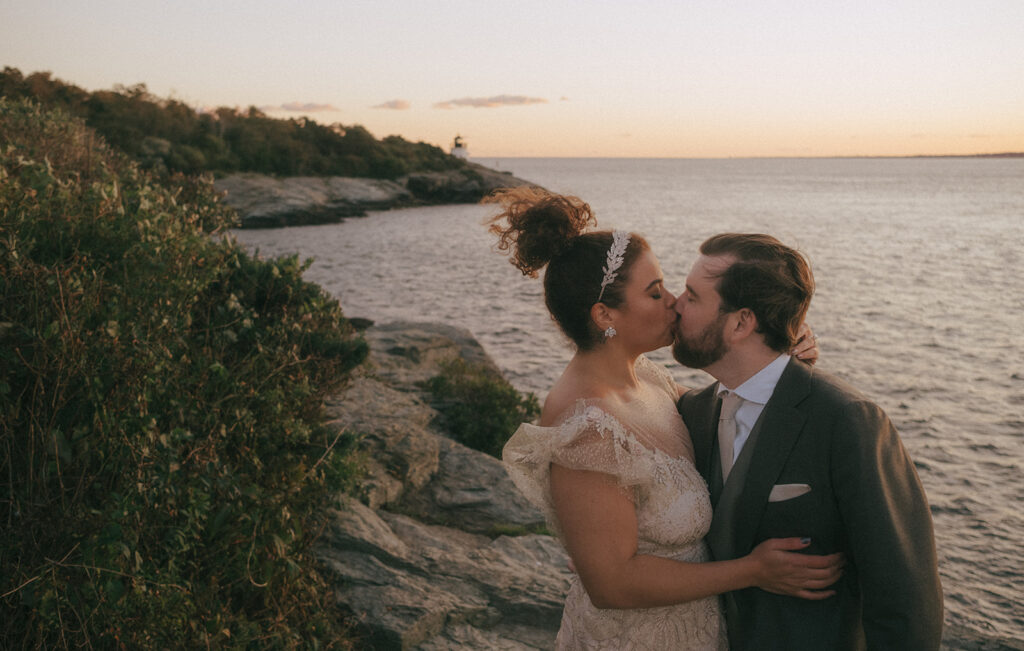 Bride and groom kiss as a breeze blows in from the bay. Castle Hill Lighthouse and the rugged coastline are picture in the background. Light is golden and pink with the setting sun. Photo by Rhode Tripper Photography