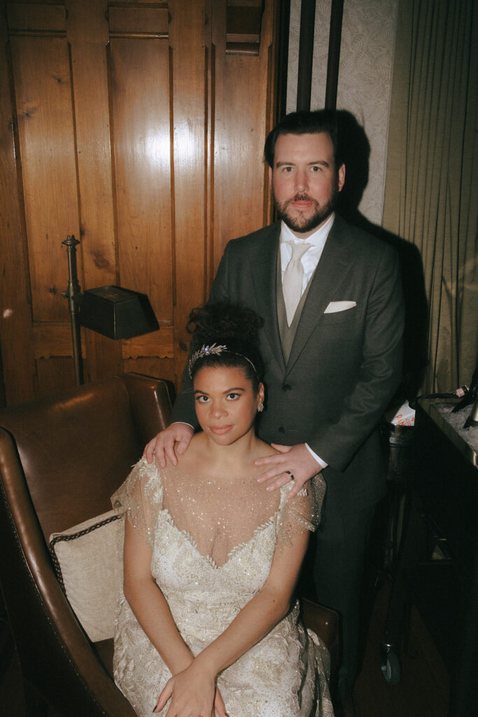 Moody, serious, direct flash photo. Bride is seated in an antique chair and groom stands behind her with his hands on her shoulders. Photo by Rhode Tripper Photography