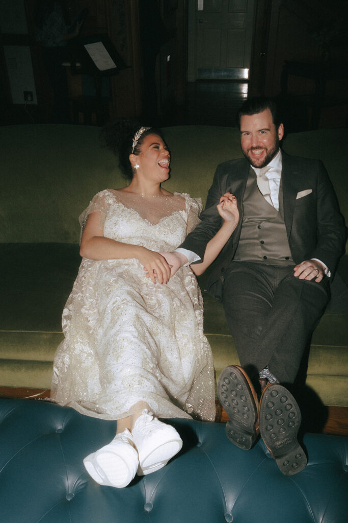 Bride and groom put their feet up on a blue leather ottoman. They are seated on a green leather coach in the lobby of Castle Hill Inn and are smiling and laughing at each other. Photo by Rhode Tripper Photography