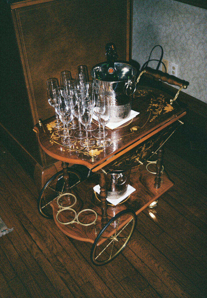 Moody flash photo of wooden antique drink cart. Cart is filled with champagne flutes and a silver ice bucket with champagne. Photo by Rhode Tripper Photography