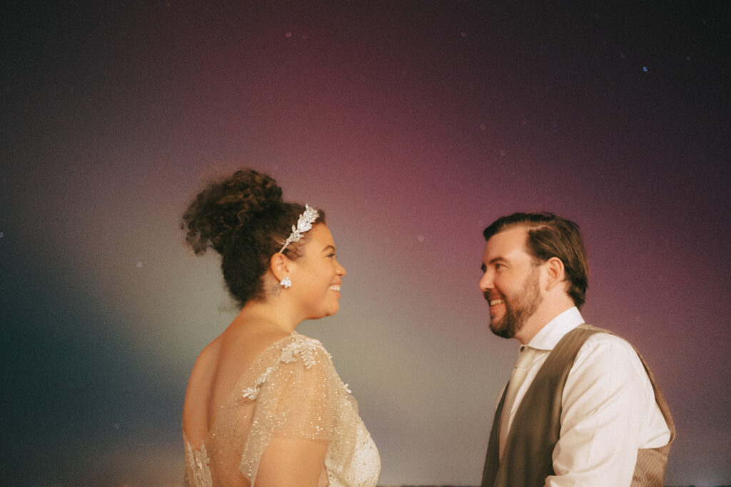 Close up of the bride and groom as they smile at each other. Colorful purple, green and red hues fill the background, showcasing the surprise Northern Lights display in Rhode Island on their wedding day at Castle Hill Inn. Photo by Rhode Tripper Photography