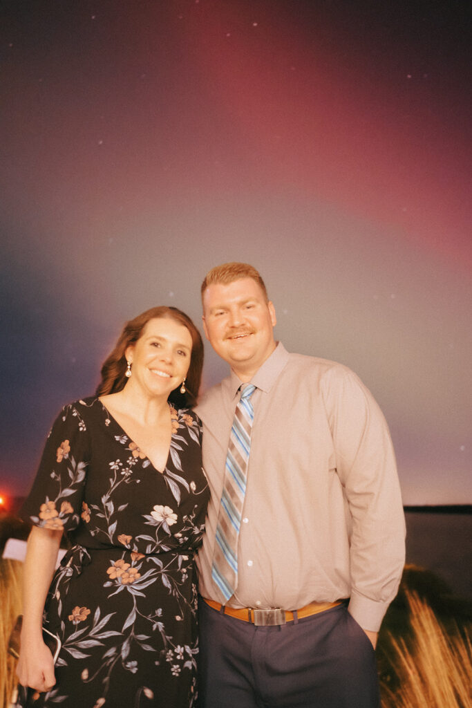 Couple stands and smiles at the camera with the Northern Lights behind them. Photo by Rhode Tripper Photography