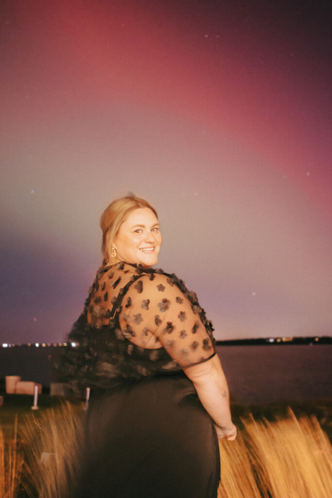 Wedding guest smiles and poses in front of the Northern Lights. Photo by Rhode Tripper Photography