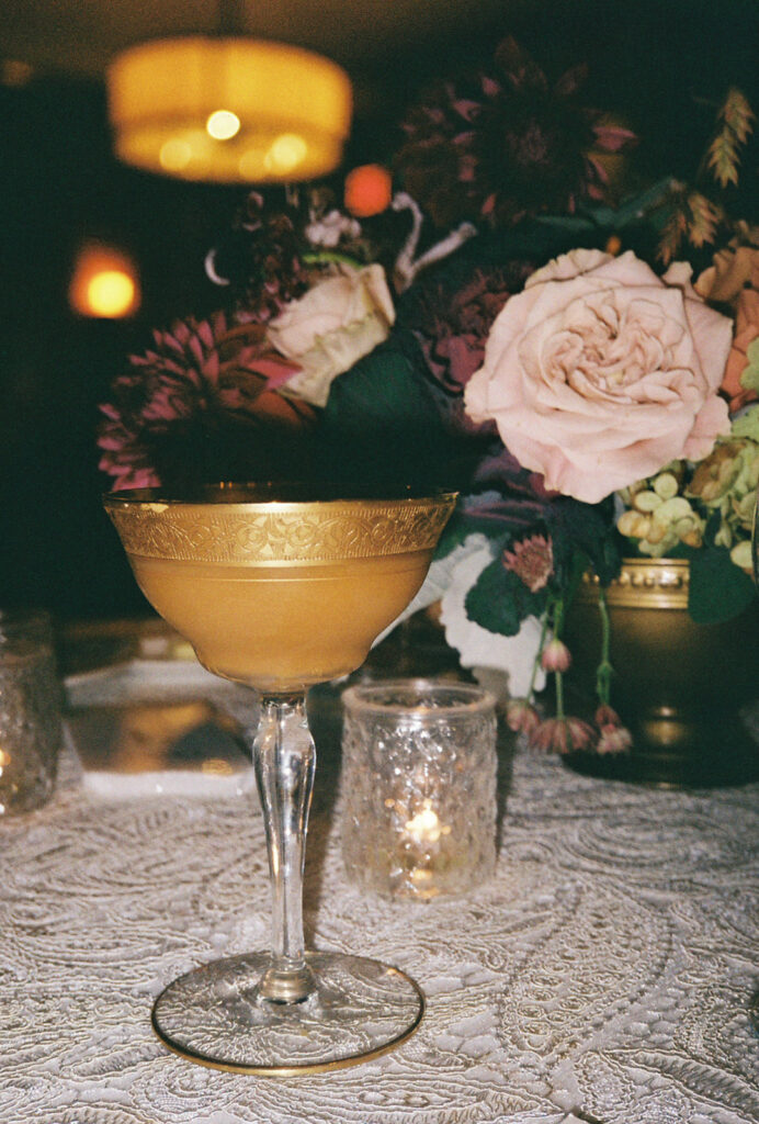 Moody film photo of an espresso martini with a gold rimmed glass. Sits atop a lace tablecloth with pink and burgundy florals behind it. Photo by Rhode Tripper Photography