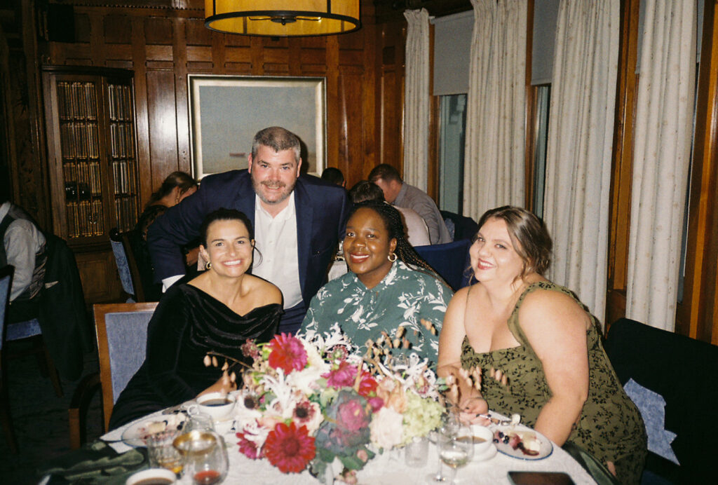 Guests Smile and look at the camera while seated and standing around their table at Castle Hill Inn wedding reception.