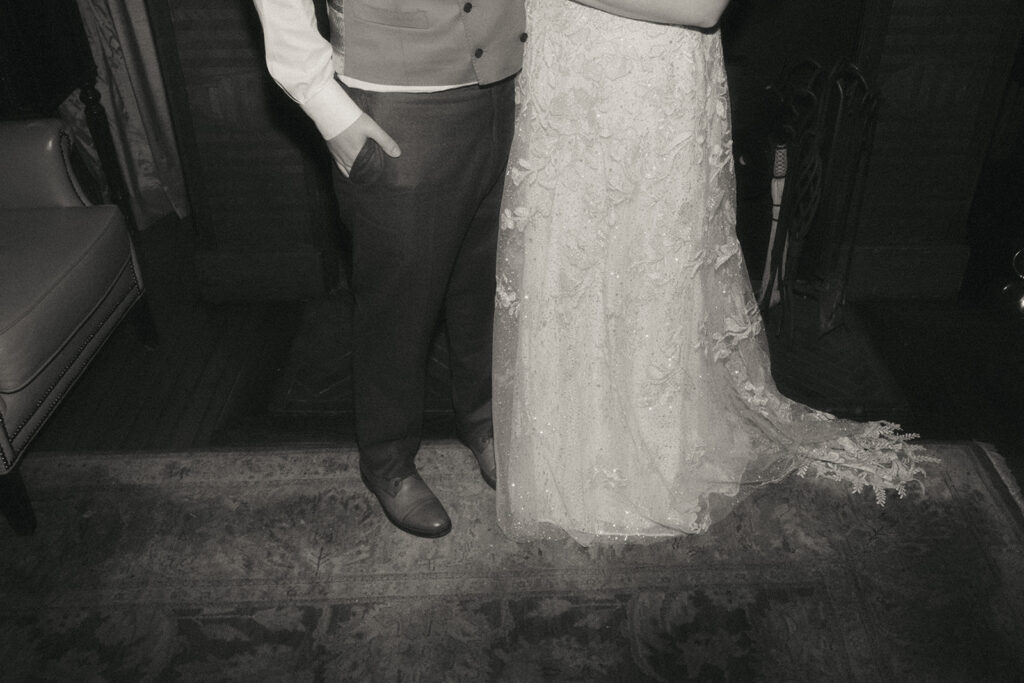 Black and white photo focuses on the lower half of the bride and groom's wedding attire - shoes, pants and dress. They stand atop a carpet in the main room of Castle Hill Inn on their wedding day. Photo by Rhode Tripper Photography