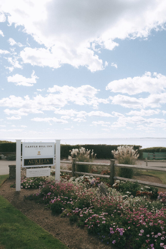 Entrance to Castle Hill Inn located on Ocean Drive in Newport, RI. A white sign and flowers sit at the bottom with the ocean and blue cloudy skies sit at the top. Photo by Rhode Tripper Photography