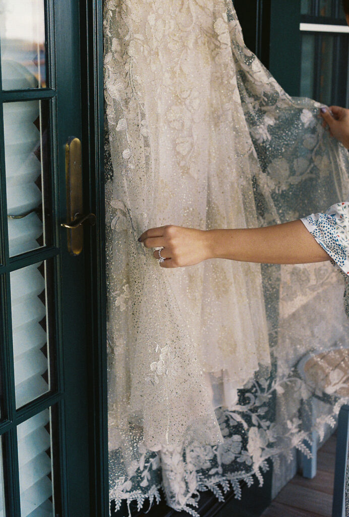 Bride holds out and admires the lace of her wedding gown. Dress is hanging up outside of her Harbor House room at Castle Hill Inn. Photo by Rhode Tripper Photography