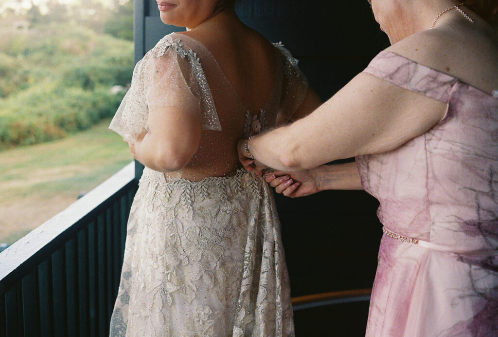 The mother of the bride helps zipper the bride's lace wedding gown. Photo is close up and located on the porch of the Harbor House at Castle Hill Inn. Photo by Rhode Tripper Photography