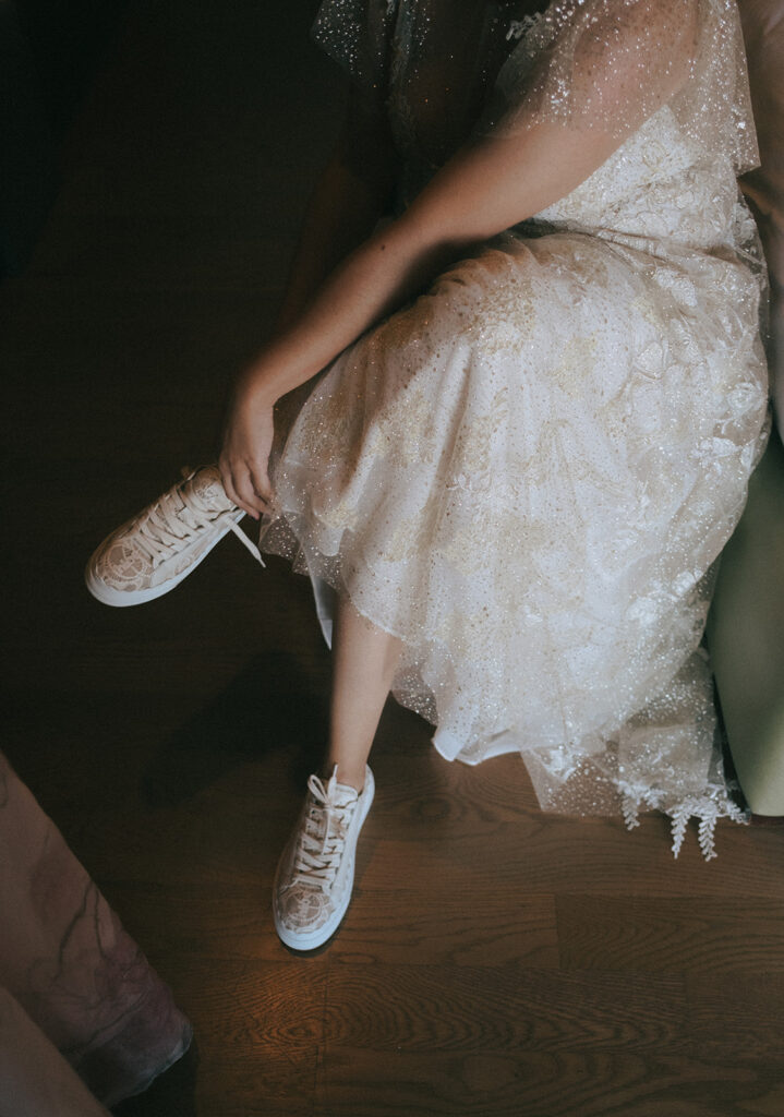 Bride crosses her legs and leans down to tie her lacy bridal sneakers while wearing her sparkly lace-covered wedding dress. Located in the Harbor House of Castle Hill Inn in Newport, RI. Photo by Rhode Tripper Photography