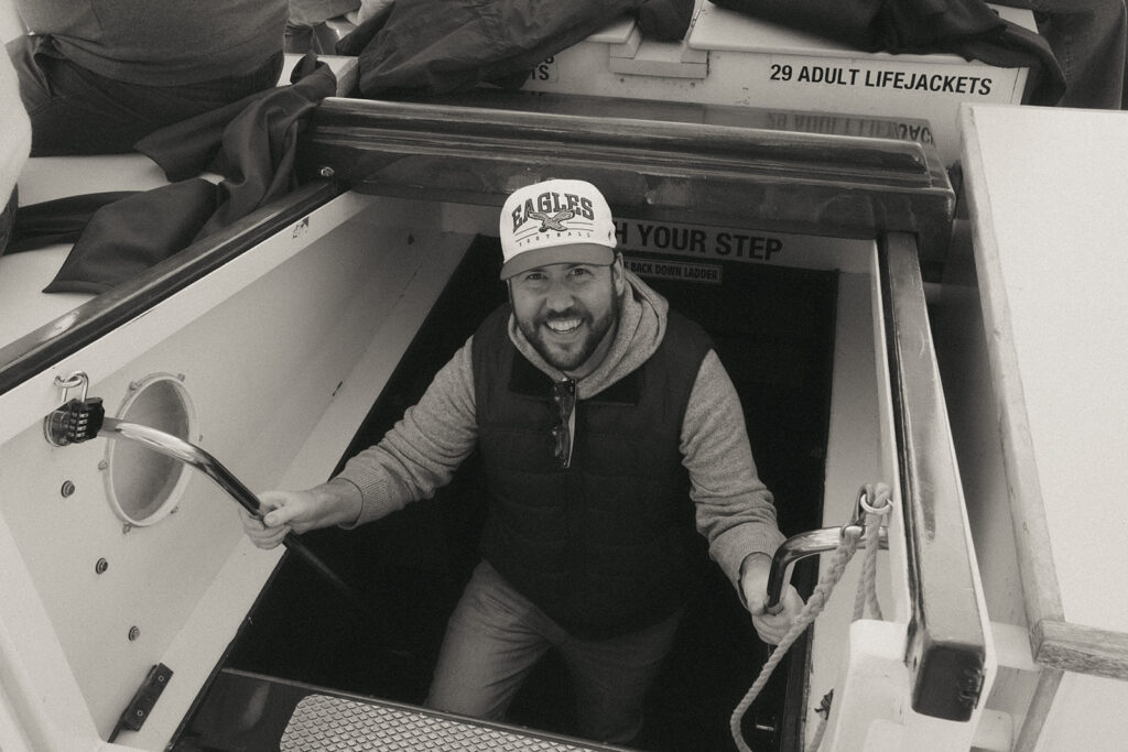 Groom in casual clothes comes out from the bottom area of the ship while sailing in Newport, RI. He has a big smile on his face. Photo by Rhode Tripper Photography