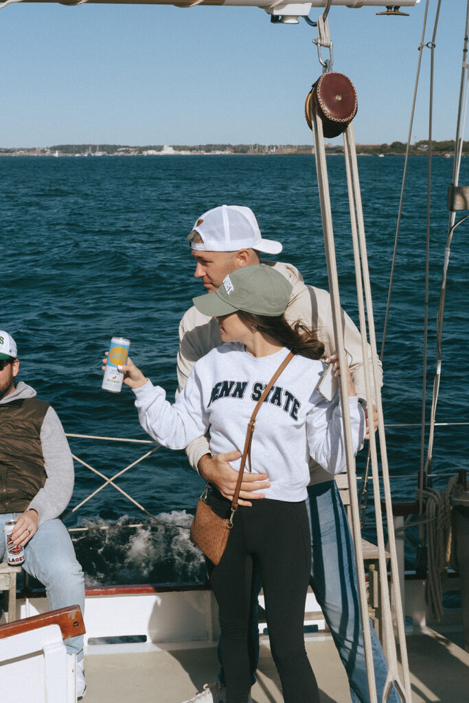 Couple holds onto each other, drinks in hand with the bay water behind them. Sailing aboard the Schooner Aquidneck in Newport. Photo by Rhode Tripper Photography