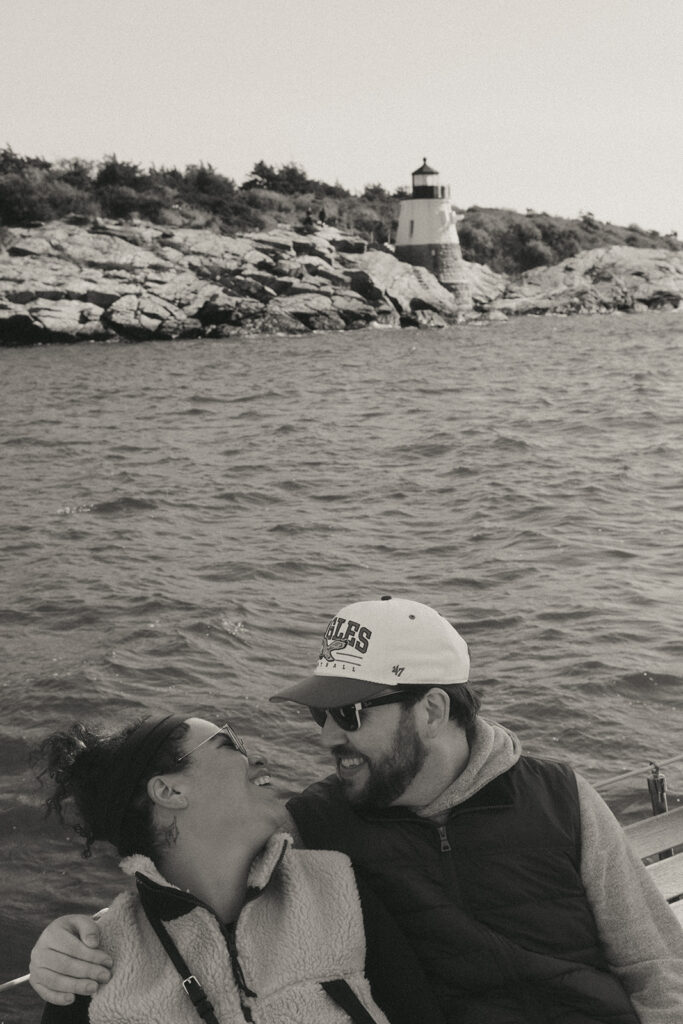 Black and white photo. Couple is seen smiling and looking into each other's eyes. The Castle Hill Lighthouse and Newport coastline is seen in the background. Photo by Rhode Tripper Photography