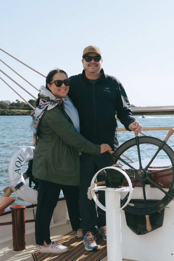 Couple hugs as they pretend to steer the sailboat. Ocean is seen behind them. Photo by Rhode Tripper Photography