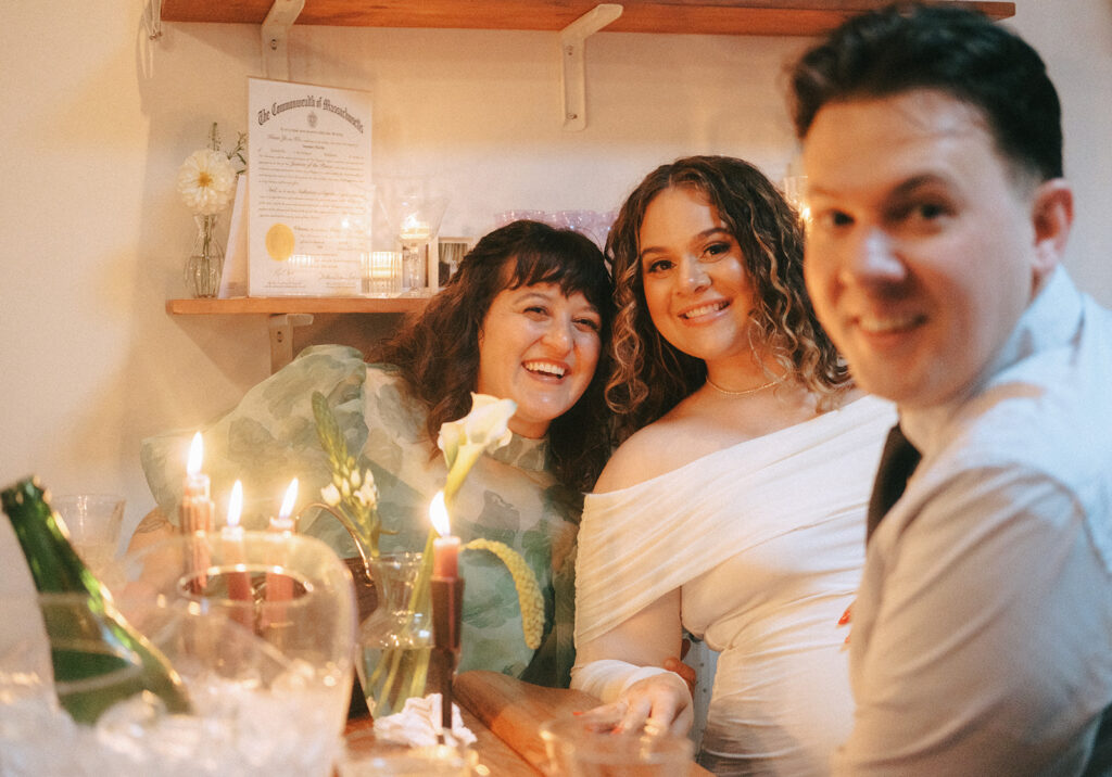 Bride smiles for the camera with her best friend and owner of Dearly Studio, Jusmine surrounded by candlelight. Jusmine's husband Blair smiles out of focus in the front right foreground of the photo.