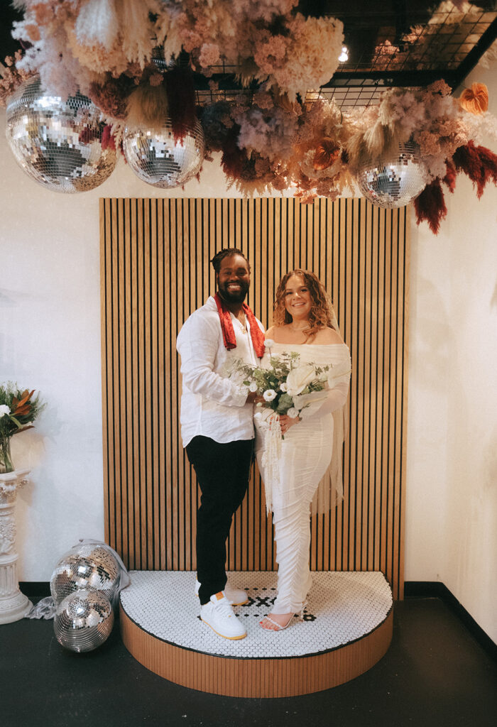 Couple smiling at camera standing on the altar at Dearly Studio in Somerville, MA. Disco balls and dried florals decorate the space.