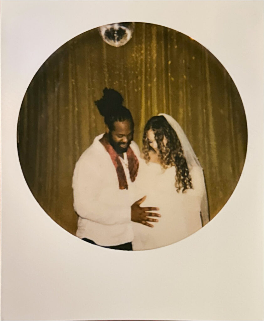 Polaroid of couple on their wedding day standing in front of a lime green velvet curtain. Husband has his hand on wife's baby bump.