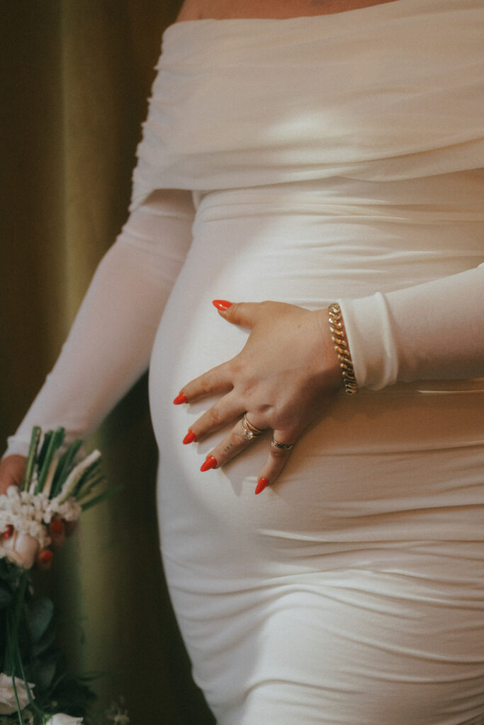 Bride holding her baby bump on her wedding day. Red wedding nails and diamond engagement ring adorn her left hand.