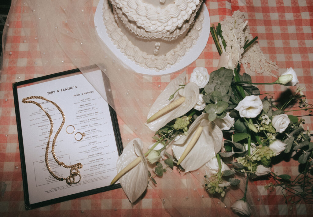 Flat lay of wedding details on a red and white checkered table cloth. Details include: Tony and Elaine's menu, bridal bouquet from Rococo Floral Co, wedding cake from Buttercream Boston, veil and wedding rings.