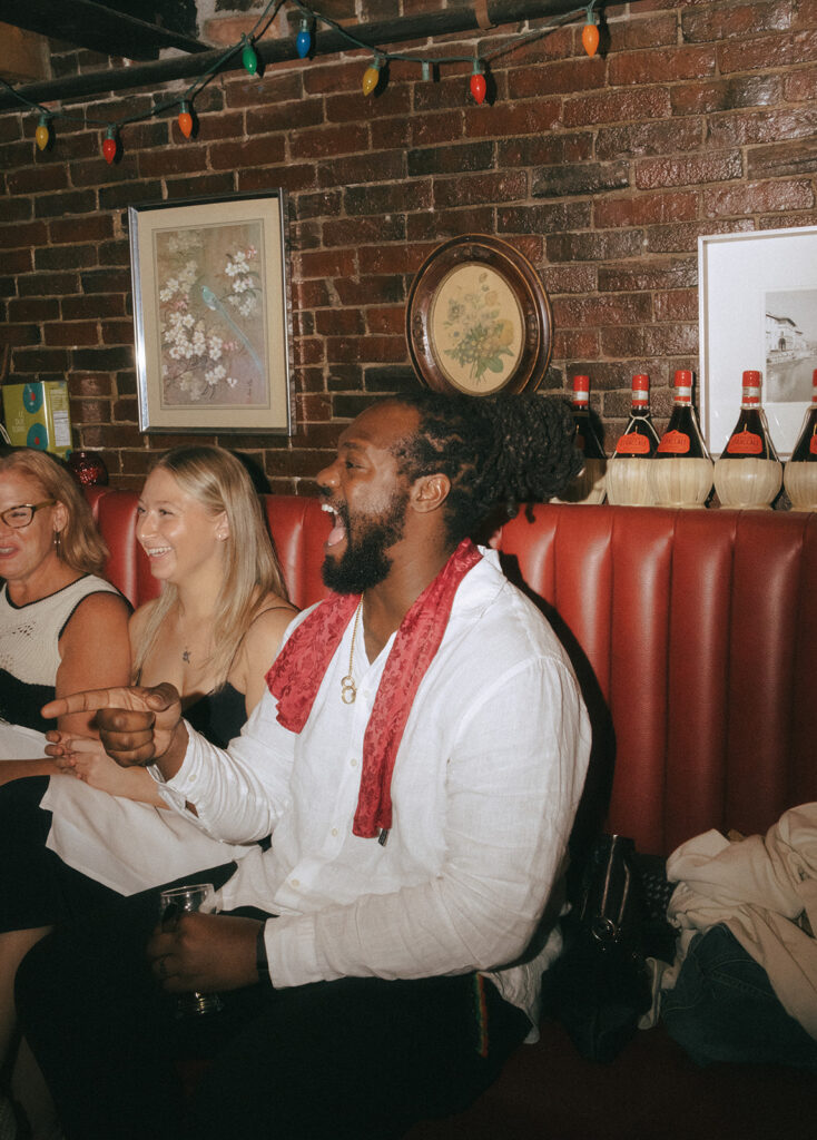 Groom laughs and smiles while conversing with his guests. Groom is seated in a red booth at Tony and Elaine's.