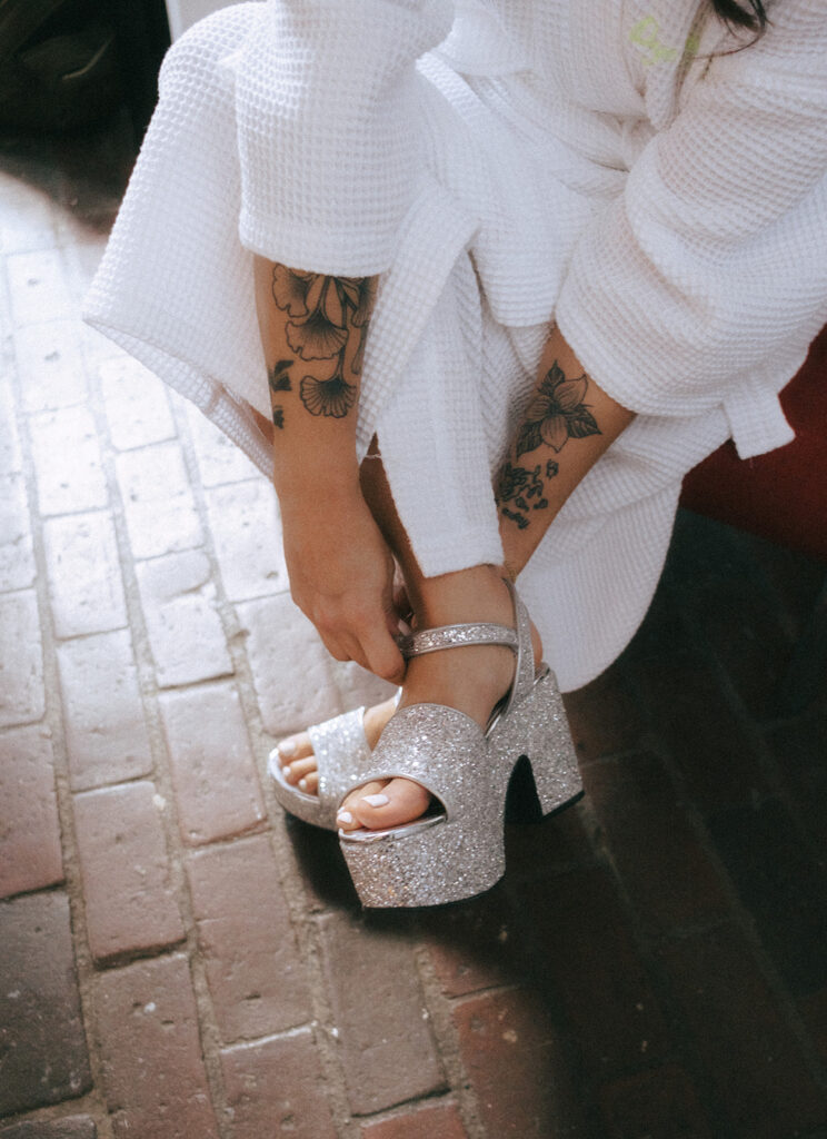 Bride buckling her sparkly platform shoes in sunlit boutique hotel room with exposed brick floors in Providence. Photo by Rhode Tripper Photography