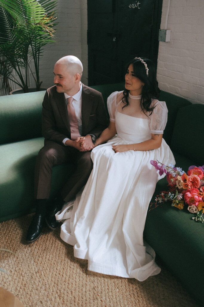 Couple bathed in natural light from large windows while seated on green velvet couch at boutique hotel spring wedding. Photo by Rhode Tripper Photography